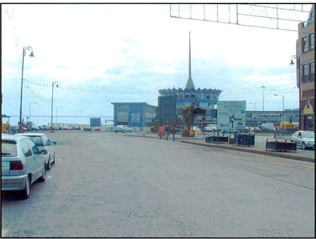 A photograph showing a wide paved harbor access area with parked cars on the left and a distinctive blue building with a spire in the background.
