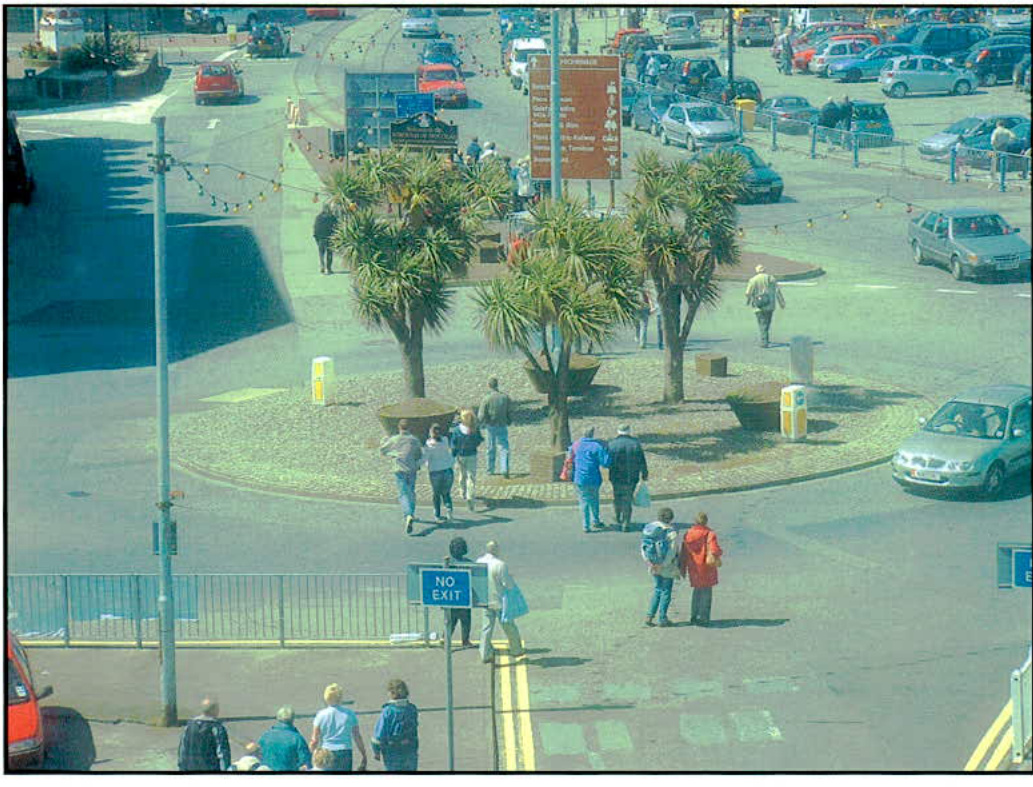 High-angle photograph of a busy roundabout featuring palm trees, pedestrians, and parked cars near a harbor area.