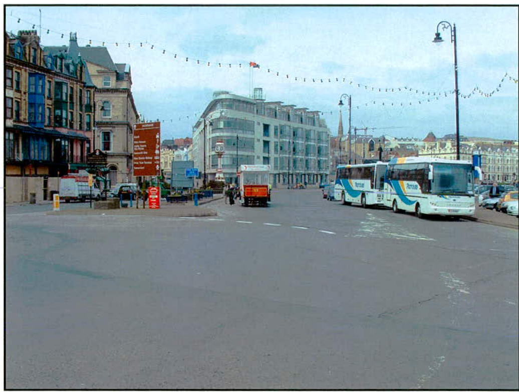 A street-level photograph showing a road junction with buses parked on the right and commercial buildings on the left, illustrating the existing highway conditions.