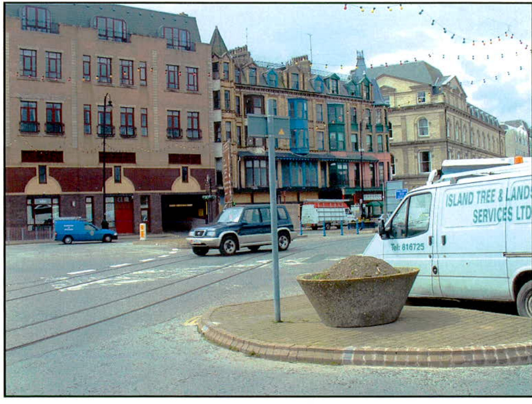 A street-level photograph showing a busy urban road with tram tracks, a roundabout with a stone planter, and a row of multi-story commercial buildings.
