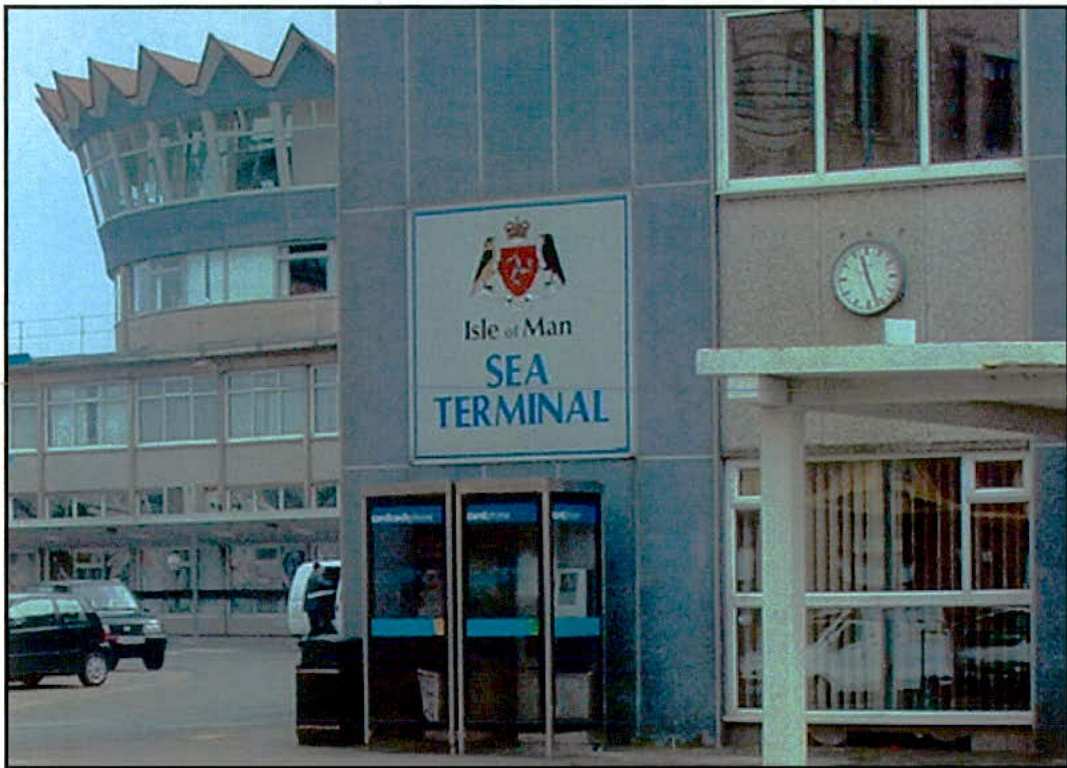 Exterior photograph of the Isle of Man Sea Terminal building featuring a sign, clock, and phone booths, with a control tower structure in the background.
