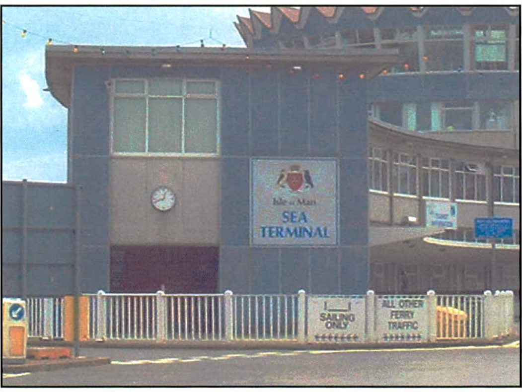 A photograph of the Isle of Man Sea Terminal building featuring a clock and signage, with a foreground fence displaying traffic instructions.