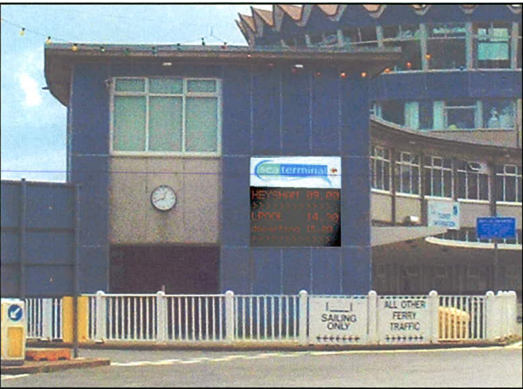 A photograph showing the exterior of a ferry terminal building with a digital departure board and a white fence in the foreground.