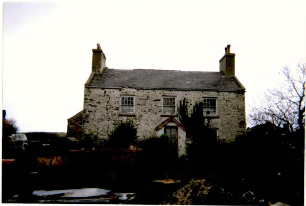 A photograph of a two-story detached stone house with a slate roof and chimneys, surrounded by vegetation.