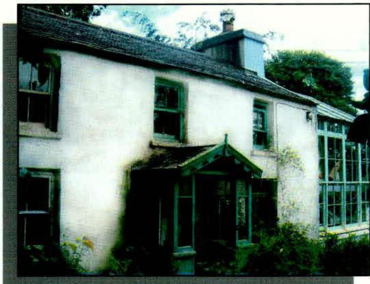 A photograph of a white-washed cottage with a slate roof and a large glass greenhouse structure attached to the side.