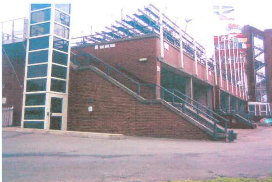 A photograph showing the exterior brickwork of a grandstand structure with external stairs and a glass elevator shaft.