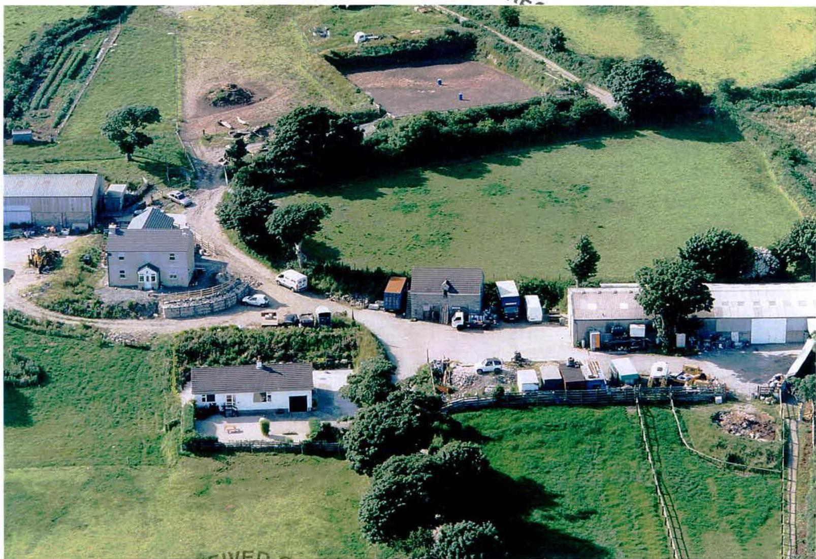 Aerial photograph of a rural farmstead showing multiple agricultural buildings, a residential house, and a cleared rectangular plot of land in a field.