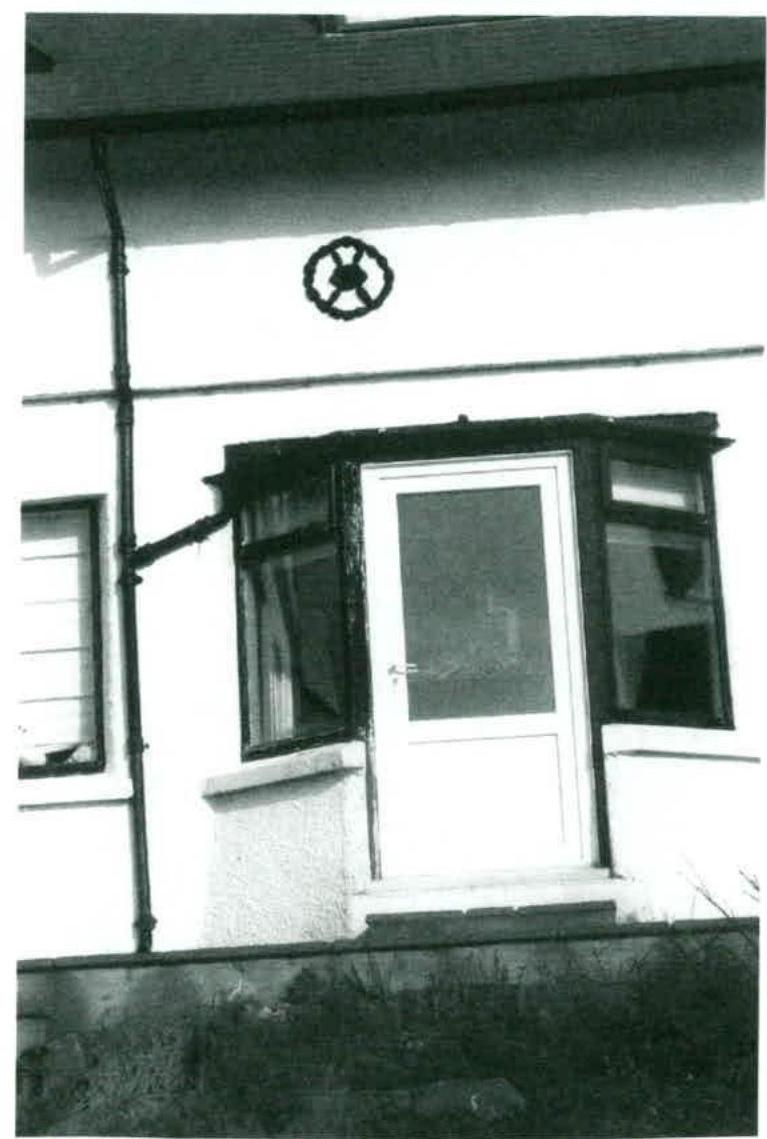 A black and white photograph showing the exterior facade of a building, featuring a central door flanked by bay windows, a drainpipe, and a decorative circular feature on the wall.