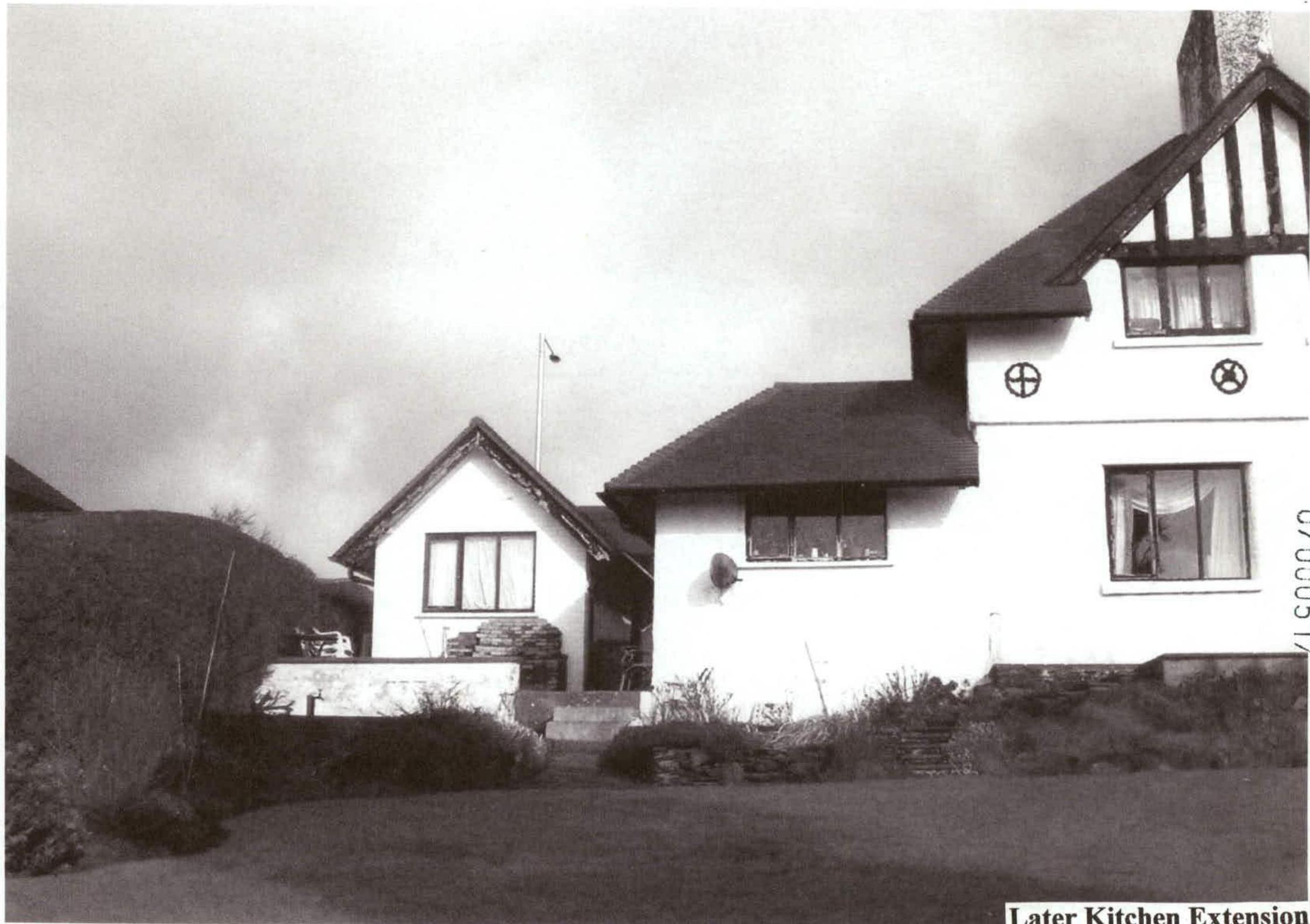 A black and white photograph showing the exterior of a detached house with a labeled kitchen extension on the left side. The image captures the seaward elevation mentioned in the application, featuring windows and a l...