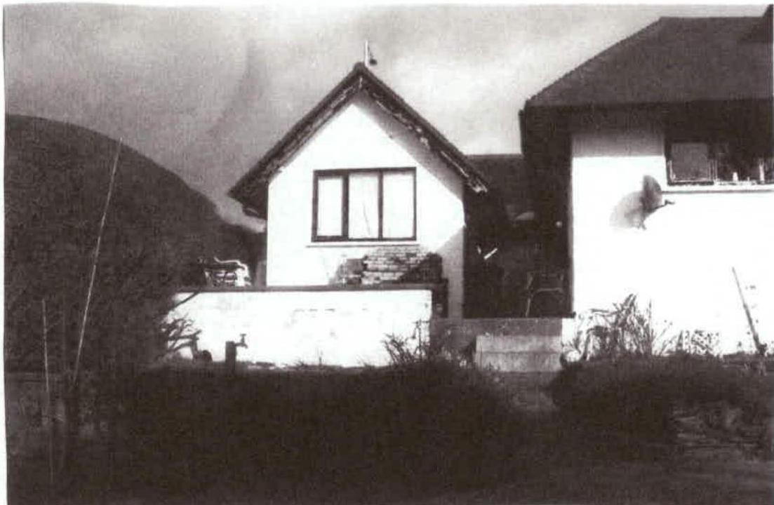 A grainy black and white photograph showing the exterior elevation of a white-walled house with a large window and a low boundary wall in the foreground.