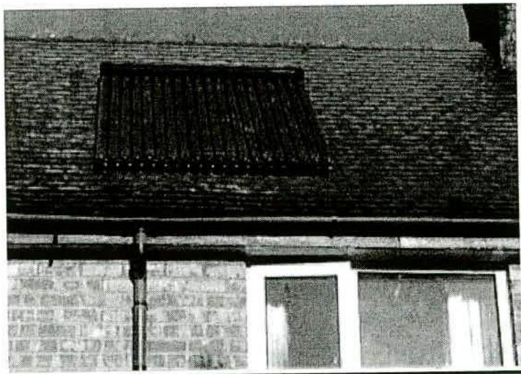 A grainy black and white photograph showing a solar thermal collector mounted on a tiled roof above a brick wall and window.