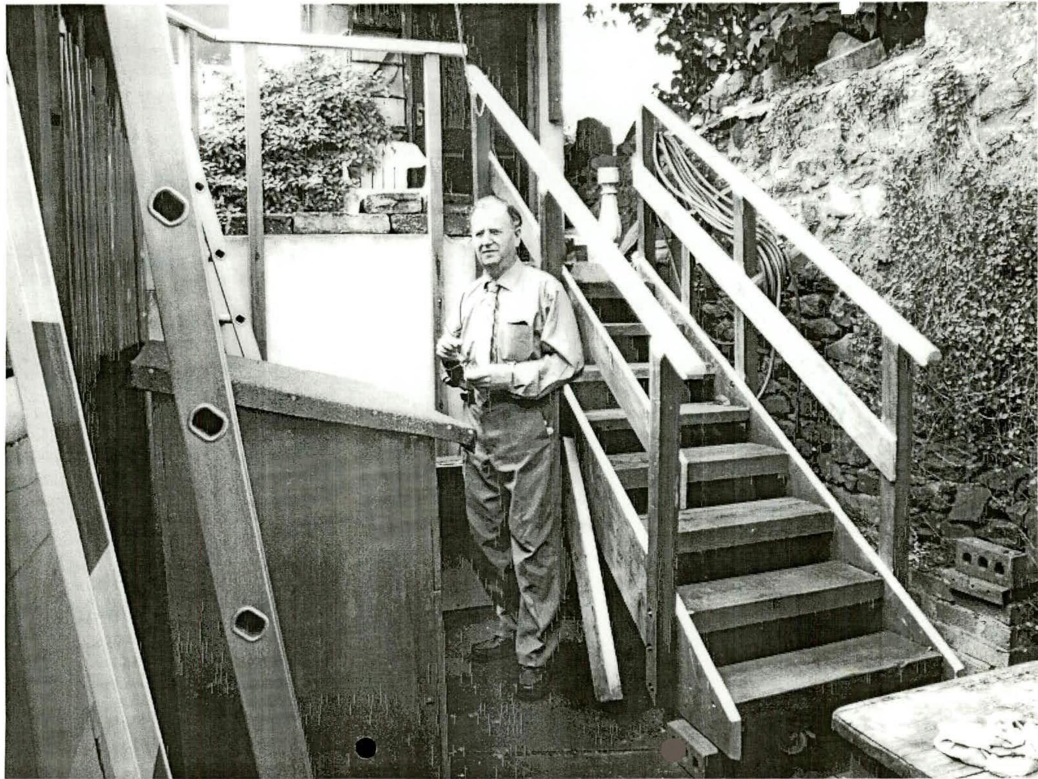 A black and white photograph showing a man standing on a wooden staircase structure under construction, with a ladder leaning nearby and a stone wall in the background.