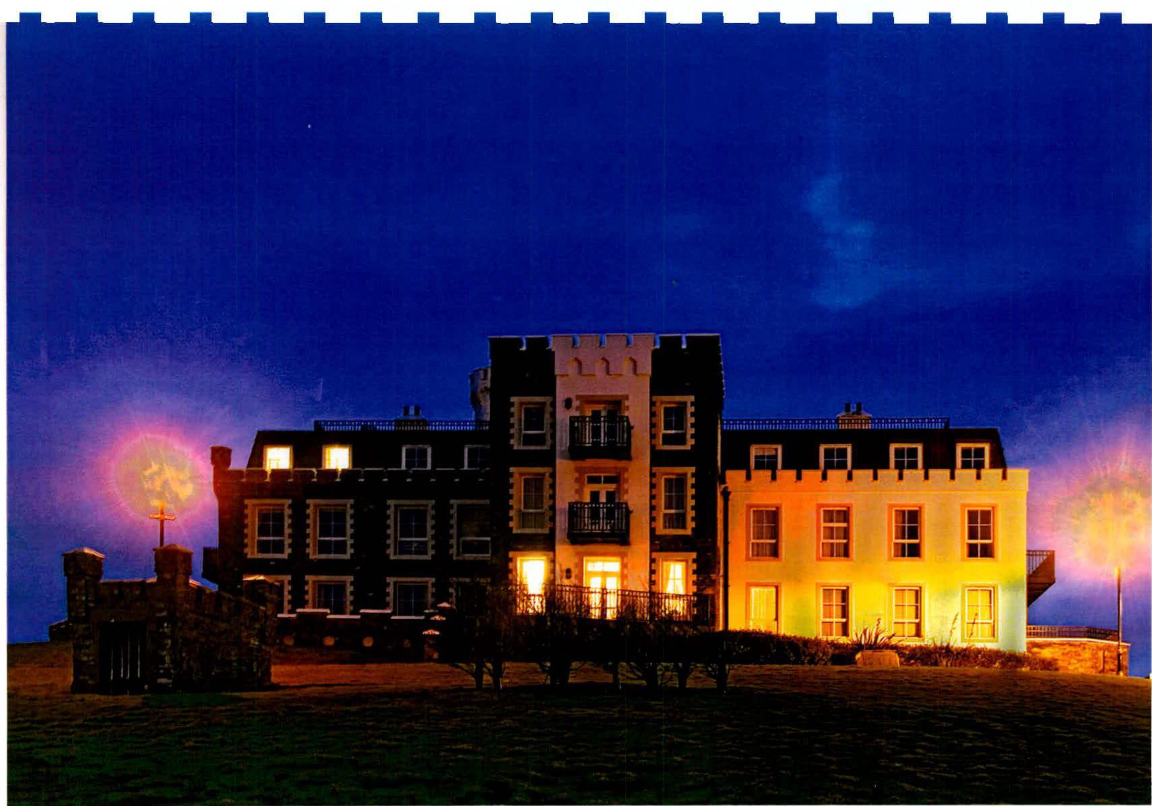 A night-time photograph of a large, multi-story detached building featuring a central crenellated tower section and balconies, illuminated against a dark blue sky.
