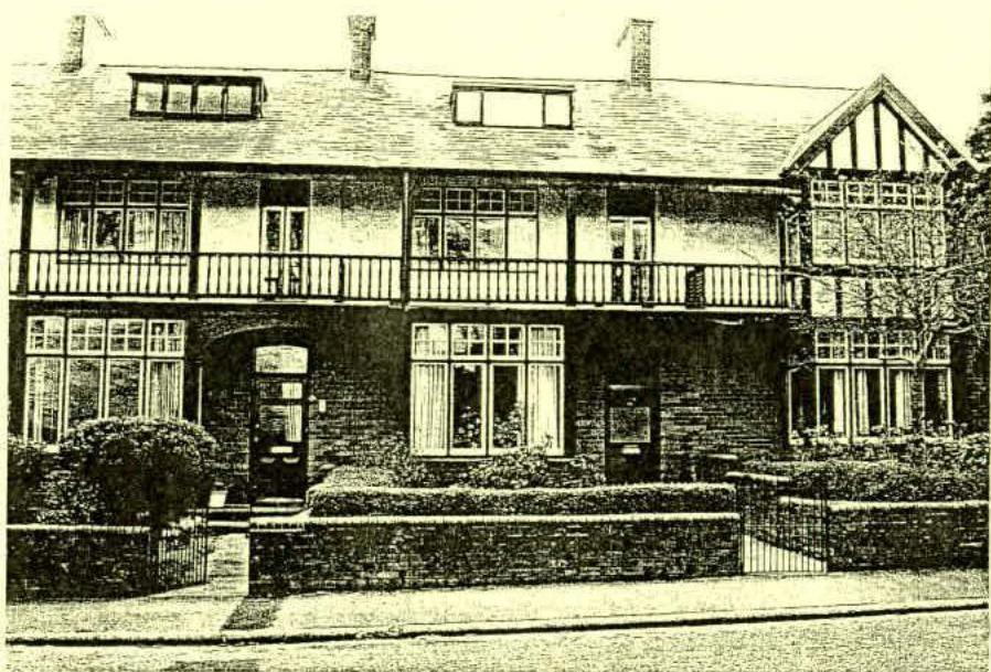 A black and white photograph showing the exterior elevation of a two-story residential building featuring a balcony, brickwork, and a boundary wall with landscaping.