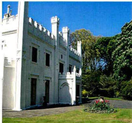 A photograph showing the exterior of a large, white building with distinctive crenellated battlements and chimneys, set against a blue sky with trees.