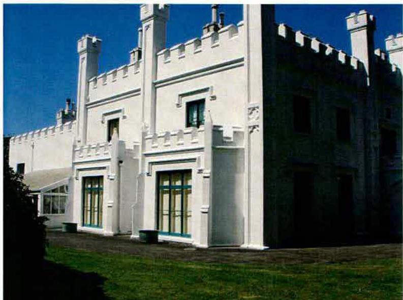 A photograph showing the exterior of a large white building with distinctive castle-like features, including crenellations and turrets, alongside a glass conservatory.