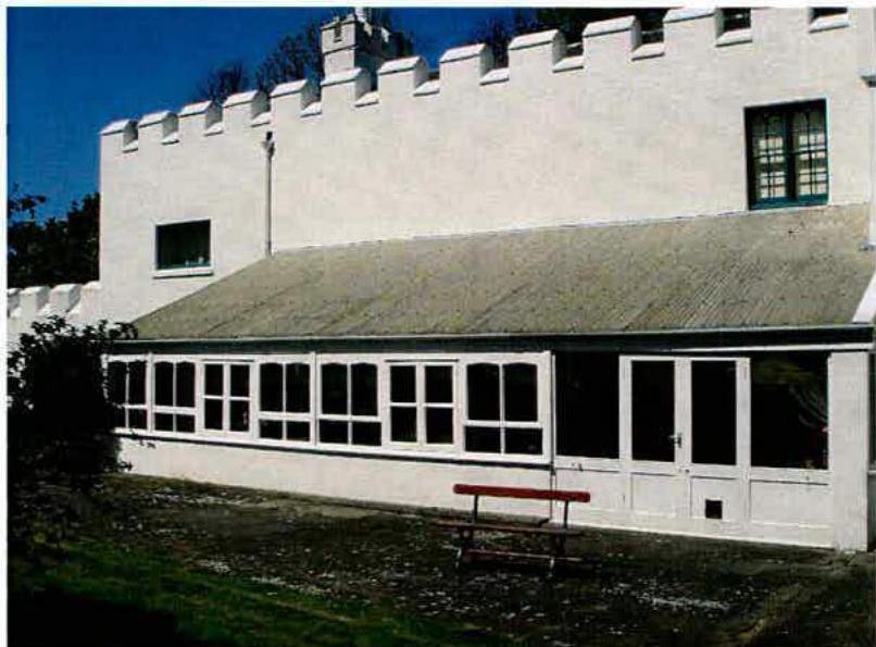 A photograph showing the exterior of a white building with a distinctive crenellated parapet and a single-story extension featuring a long row of windows.