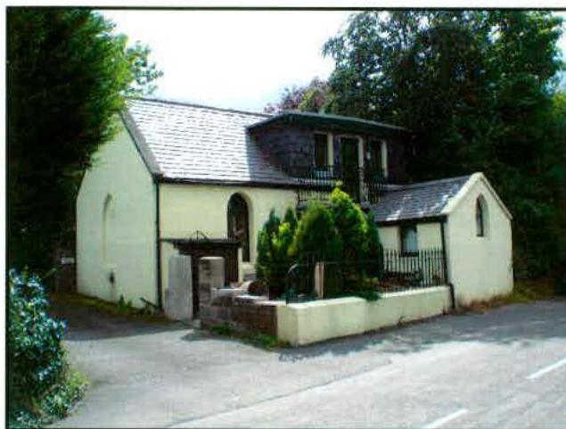 A photograph of a cream-colored detached house with a slate roof, featuring a dark stone upper section with a balcony and a smaller attached wing.