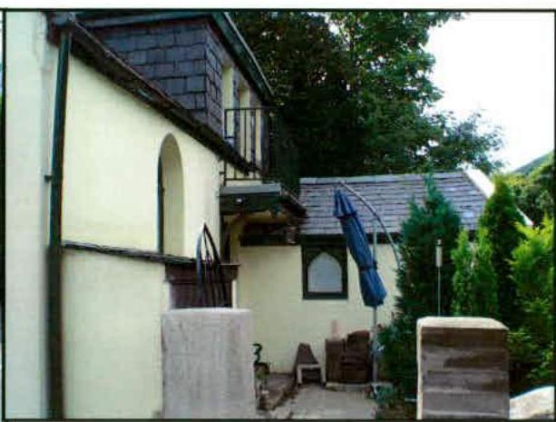 A photograph showing the rear exterior of a cream-colored house with half-timbered details, featuring a balcony and a patio area with a closed umbrella.