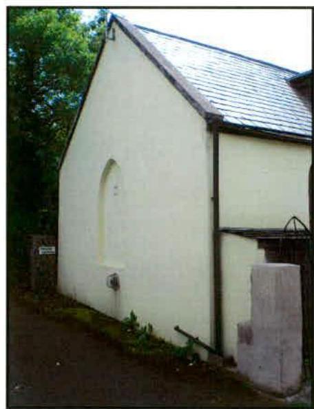 A photograph showing the exterior side elevation of a white-walled building with a slate roof and an arched window niche.