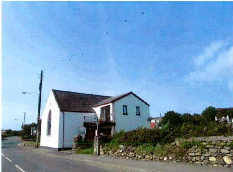 A photograph showing a white building, likely a converted chapel, with a modern extension featuring a balcony and roof terrace situated by a roadside.