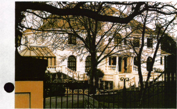 A grainy photograph showing a large, white detached house partially obscured by bare tree branches and a foreground fence.