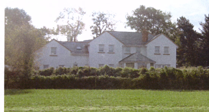 A grainy photograph showing a large white two-story building with a slate roof, surrounded by mature trees and a hedge in a rural setting.