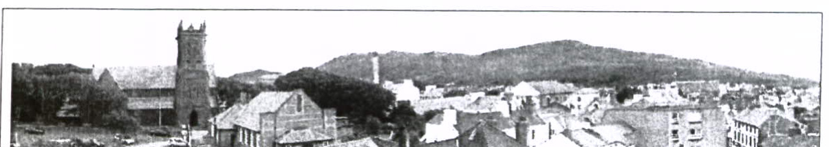 A black and white panoramic photograph showing a town scene with a prominent church tower and a large hill in the background.