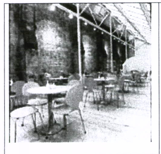 Grainy black and white photograph showing the interior of a large building with stone walls, high ceilings, and tables and chairs.