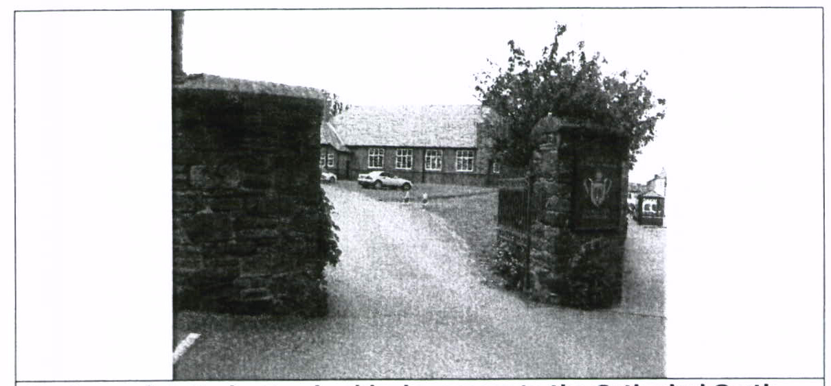 A grainy black and white photograph showing a driveway entrance flanked by stone pillars, leading towards a large building with parked cars in the background.