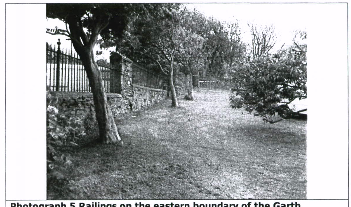 A black and white photograph showing a grassy area with trees and a stone wall topped with iron railings along the left side. The caption identifies this as the eastern boundary of the property.