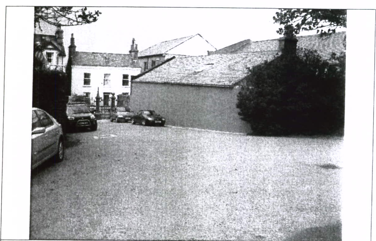 A grainy black and white photograph showing a street scene with parked cars and existing buildings, including a house and a long single-story structure.