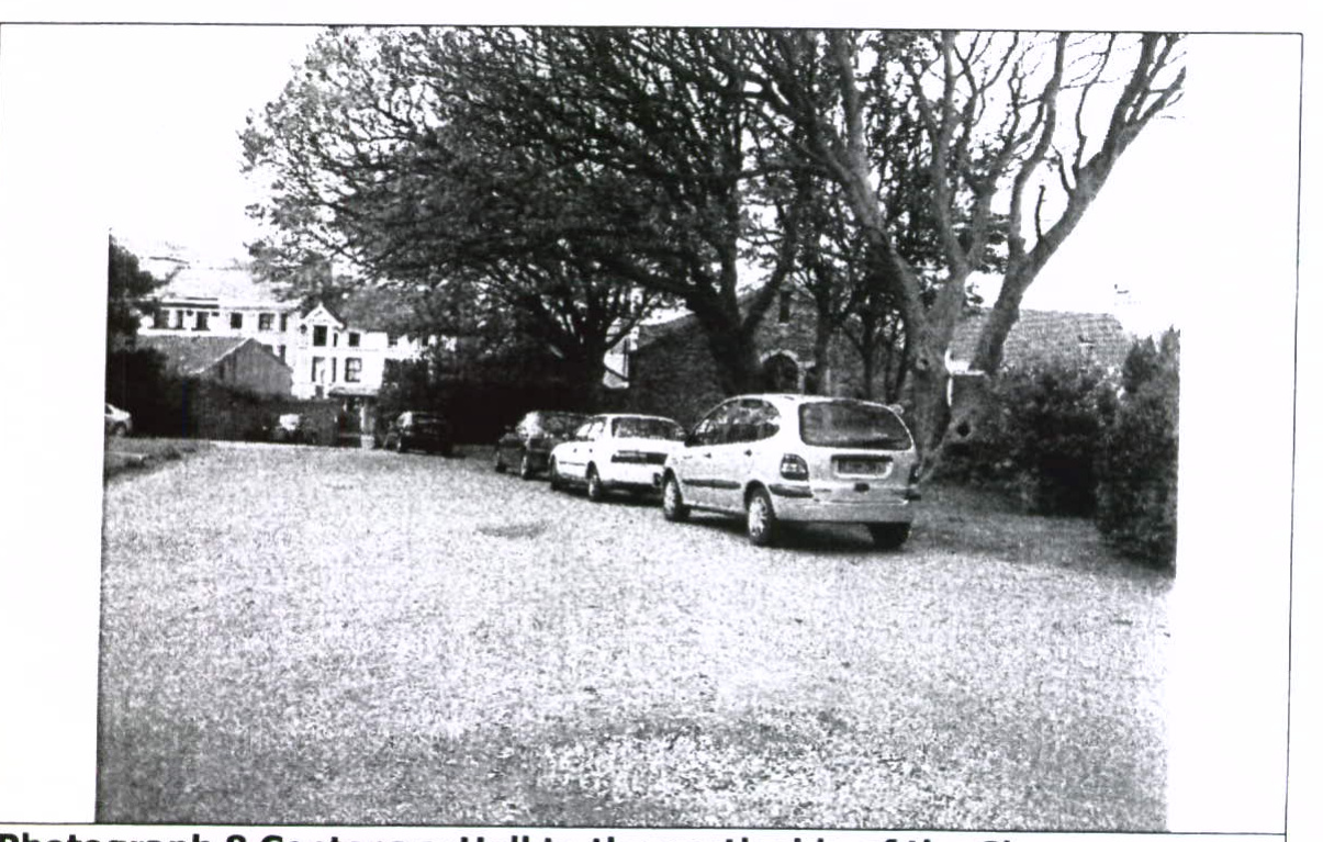 A black and white photograph showing a gravel driveway with several parked cars and large trees, with buildings visible in the background.