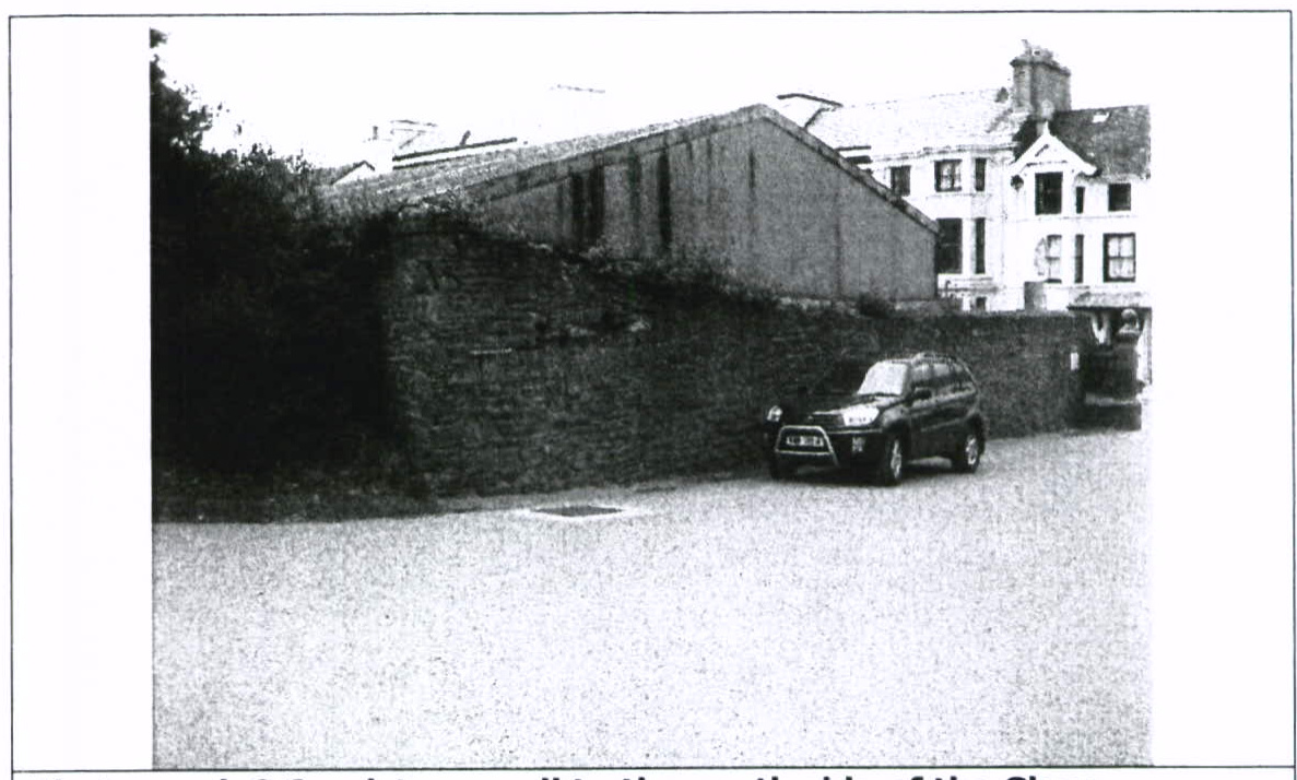 A grainy black and white photograph showing a stone boundary wall with a long, low barn-like building behind it and a larger white house in the background.