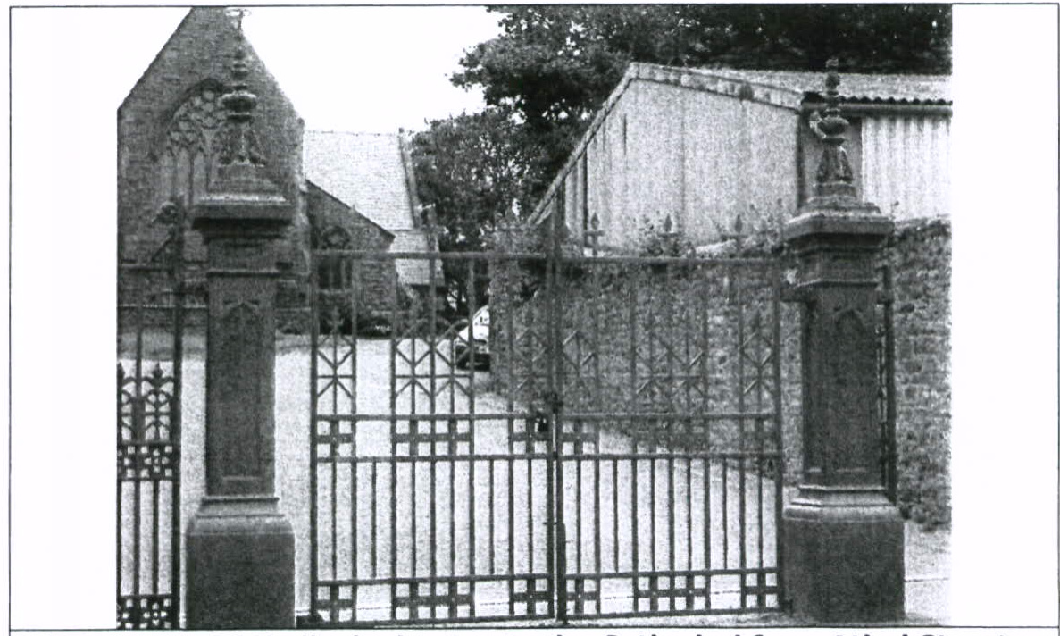 A black and white photograph showing an ornate metal gate flanked by stone pillars at the entrance to a property. Behind the gate, a driveway leads to stone buildings, including a structure with a pointed gable and a...