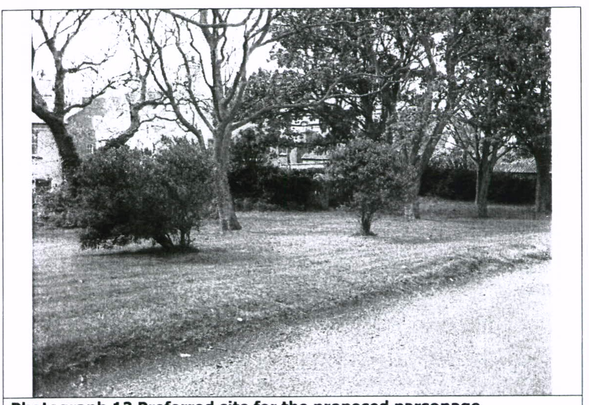 A black and white photograph showing a grassy plot of land with mature trees and shrubs, with buildings visible in the background.