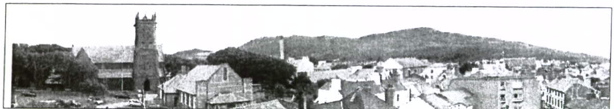 A black and white panoramic photograph depicting a townscape with a prominent church tower on the left and hills in the distance.