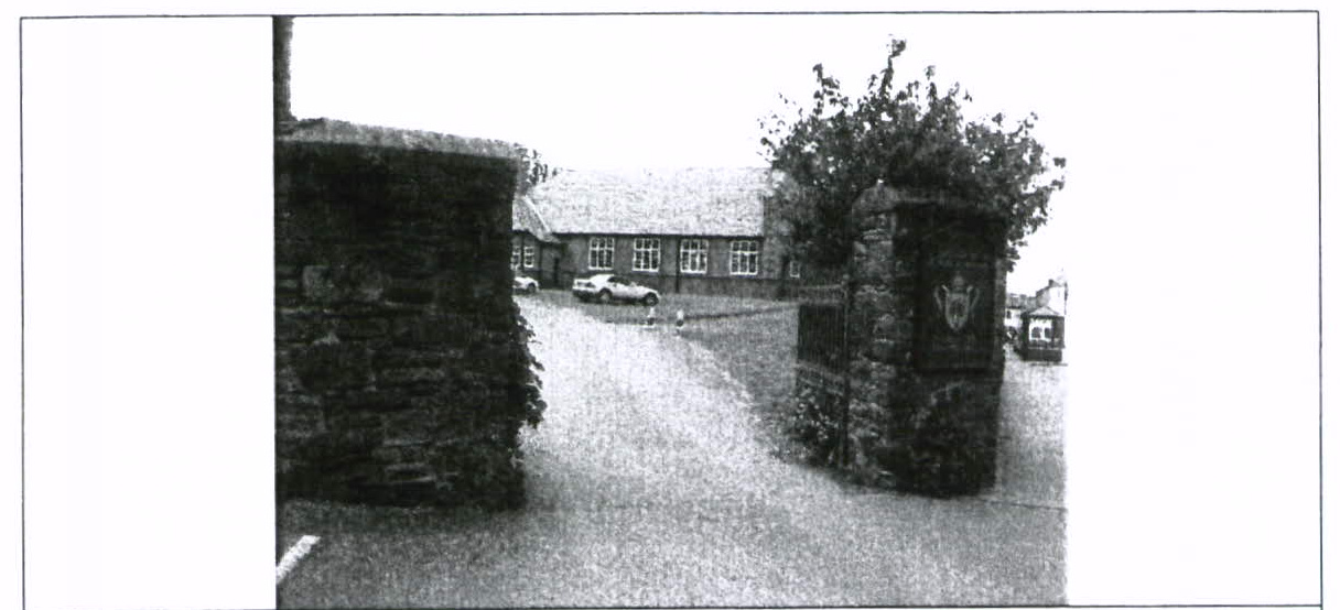 A black and white photograph showing a stone entrance gate with pillars and a long, single-story building in the background with cars parked in front.