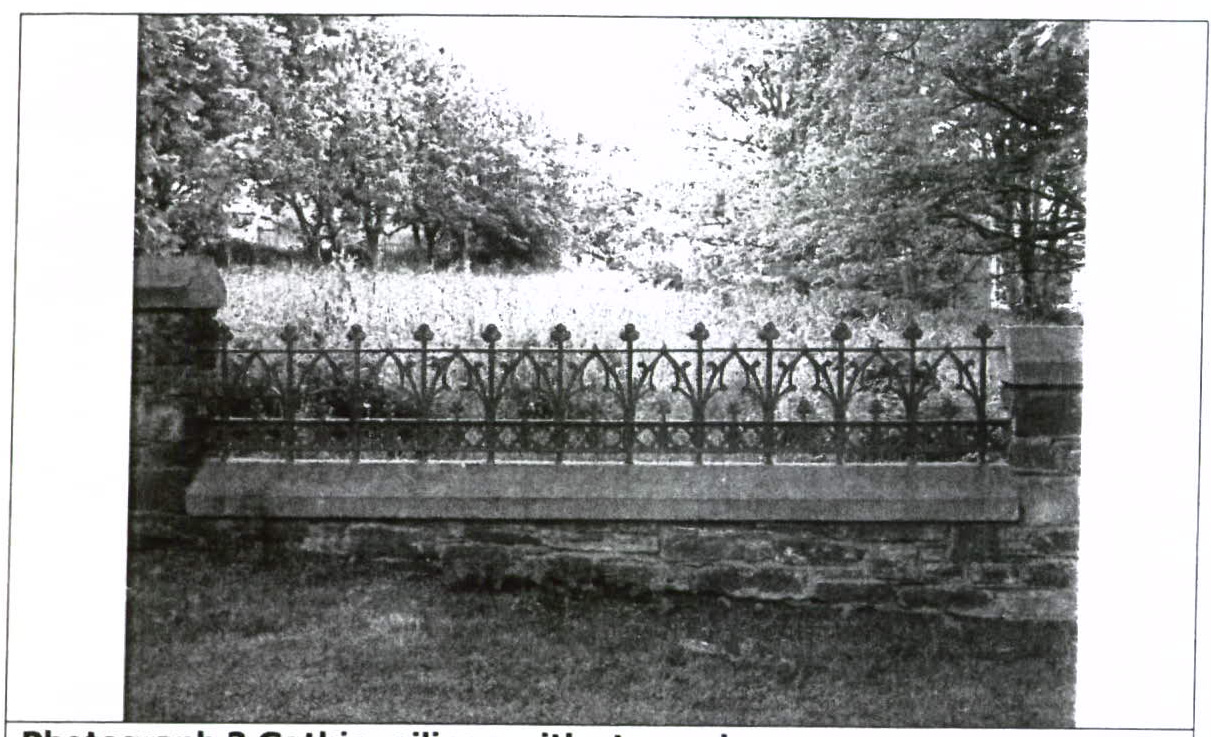 A black and white photograph showing a stone wall topped with an ornate metal railing featuring Gothic-style pointed arches, set against a backdrop of trees and grass.