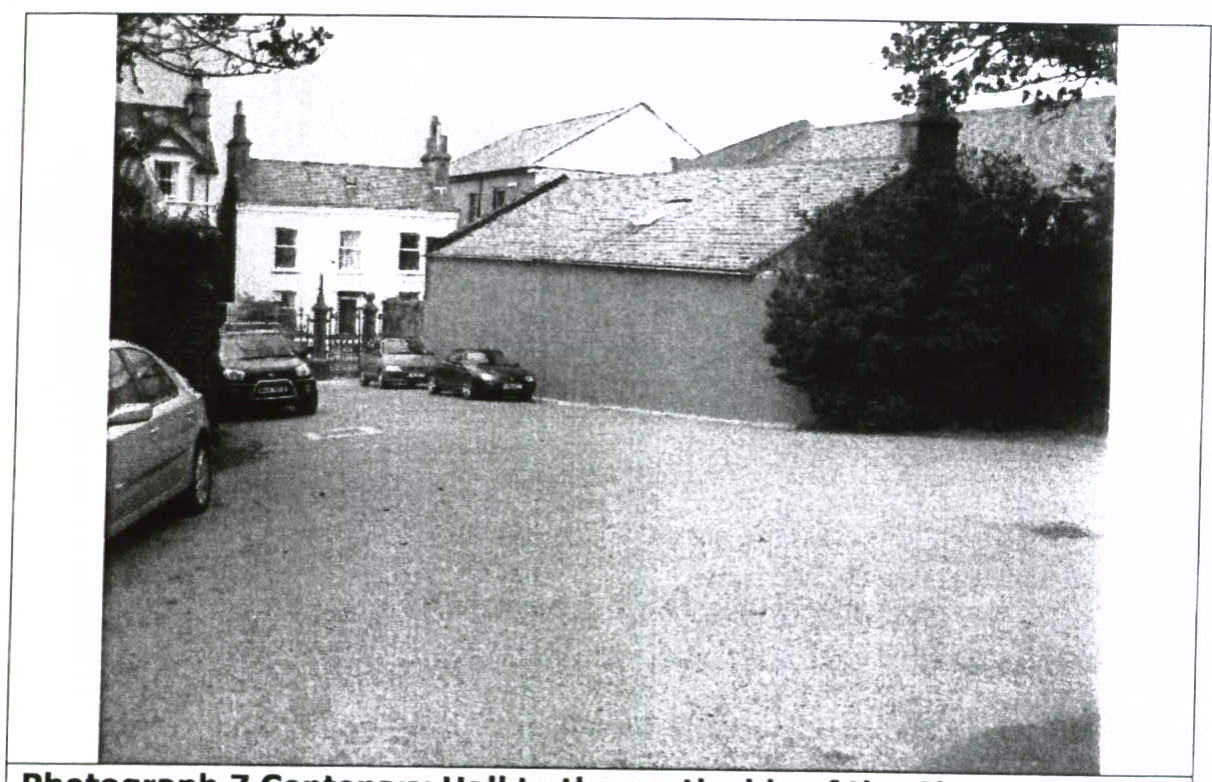 A grainy black and white photograph showing a paved area with parked cars and traditional buildings with slate roofs in the background.