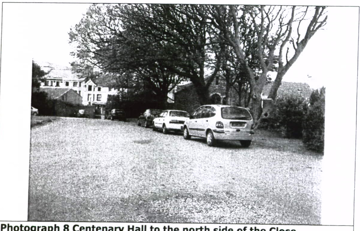 A black and white photograph showing a gravel driveway with several parked cars in the foreground. In the background, large trees and buildings are visible, including a white house and a stone structure identified by...