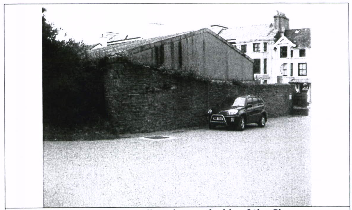A black and white photograph showing a long stone-walled outbuilding or barn behind a boundary wall, with a larger white house visible to the right and a car parked in the foreground.