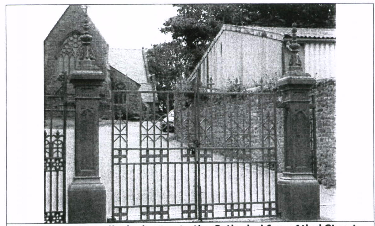 A black and white photograph showing an ornate metal gate with stone pillars, leading to a property with stone buildings and a barn-like structure in the background.