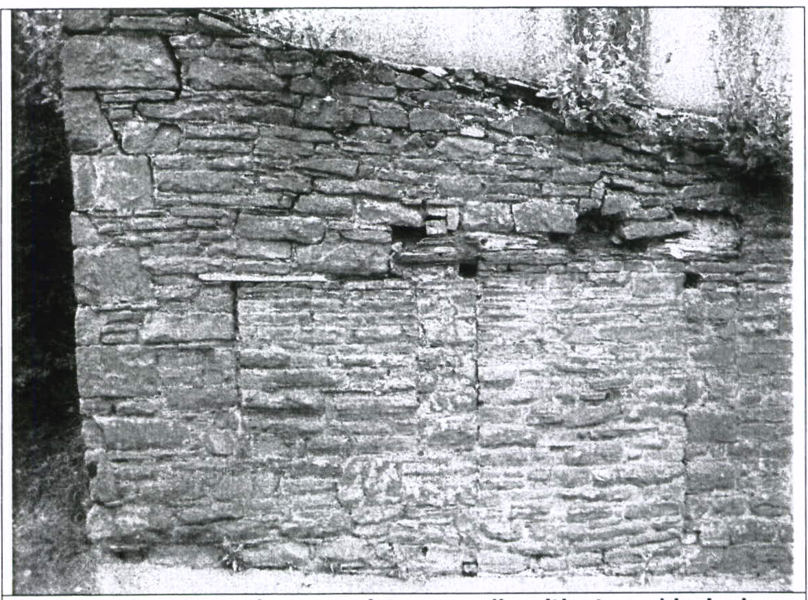 A black and white photograph showing a close-up of a weathered stone wall with rough masonry and vegetation growing along the top edge.