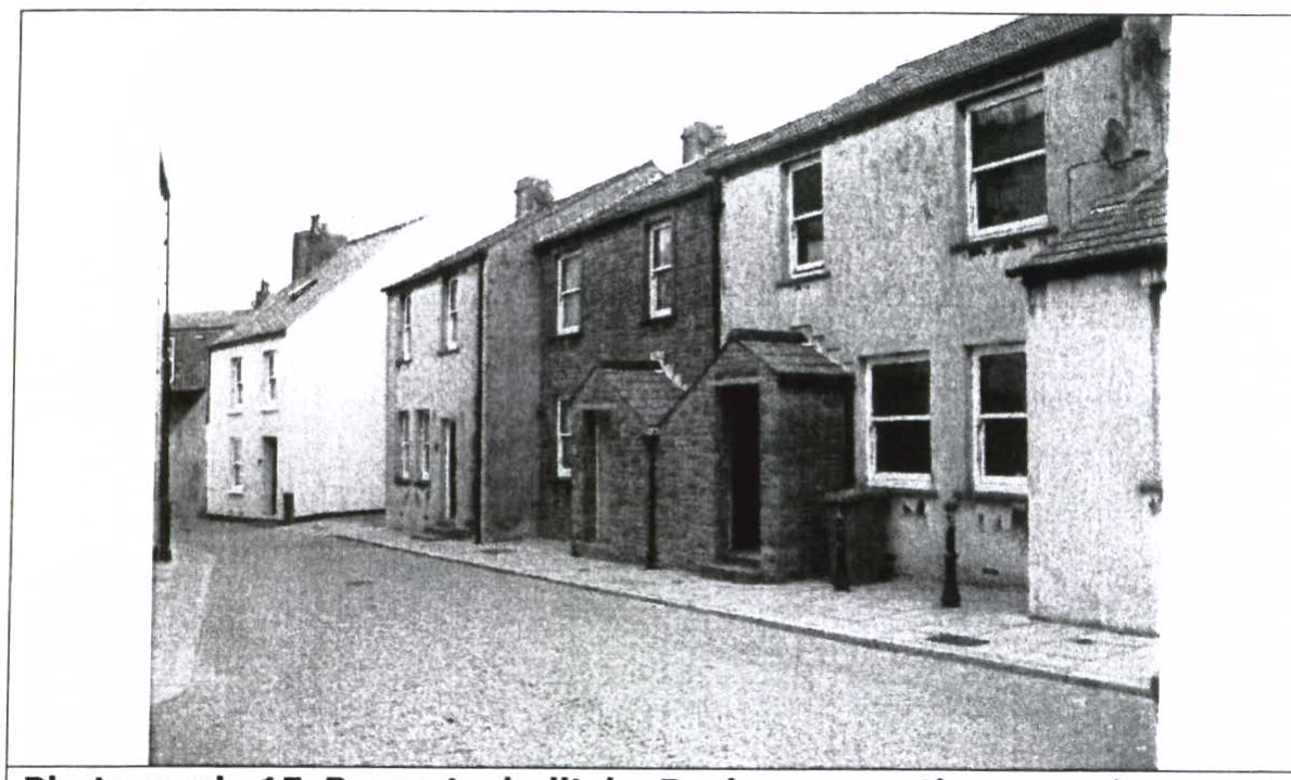 A grainy black and white photograph showing a row of terraced buildings along a street, likely depicting the existing site context.