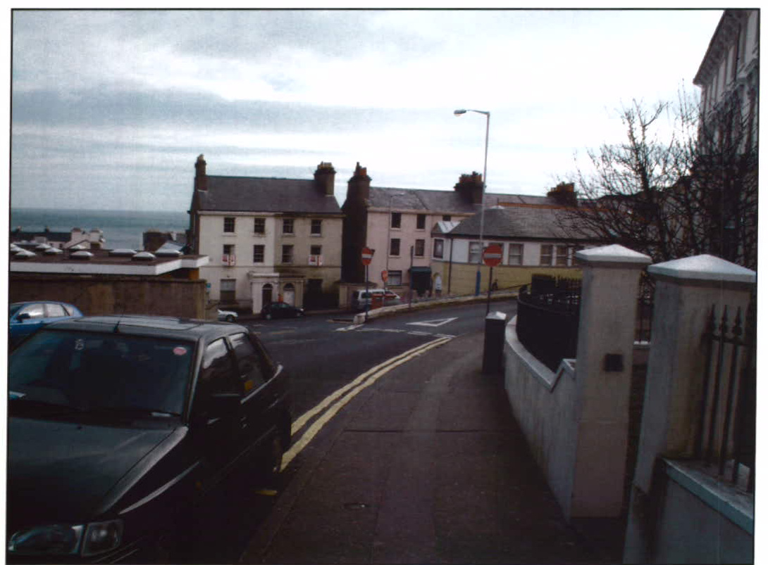 A street-level photograph showing a row of buildings near the coast with parked cars and a boundary wall in the foreground.