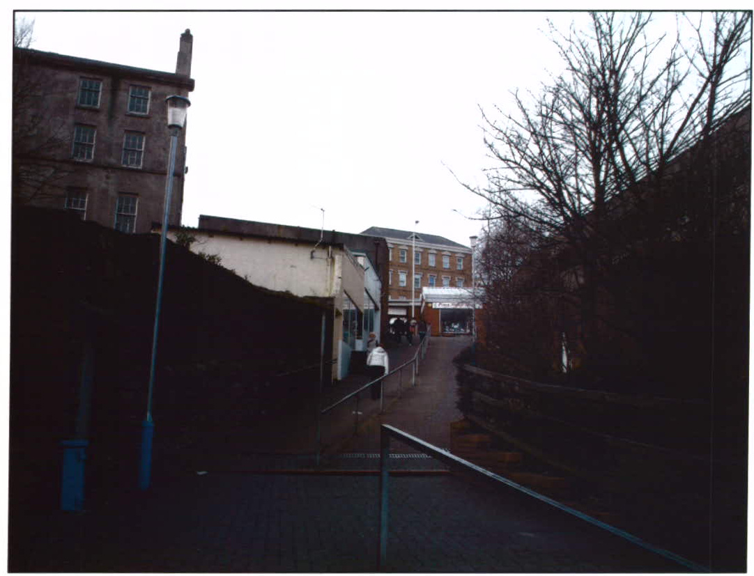 A street-level photograph showing a paved pedestrian path leading past a large multi-story building on the left and lower commercial structures in the background.