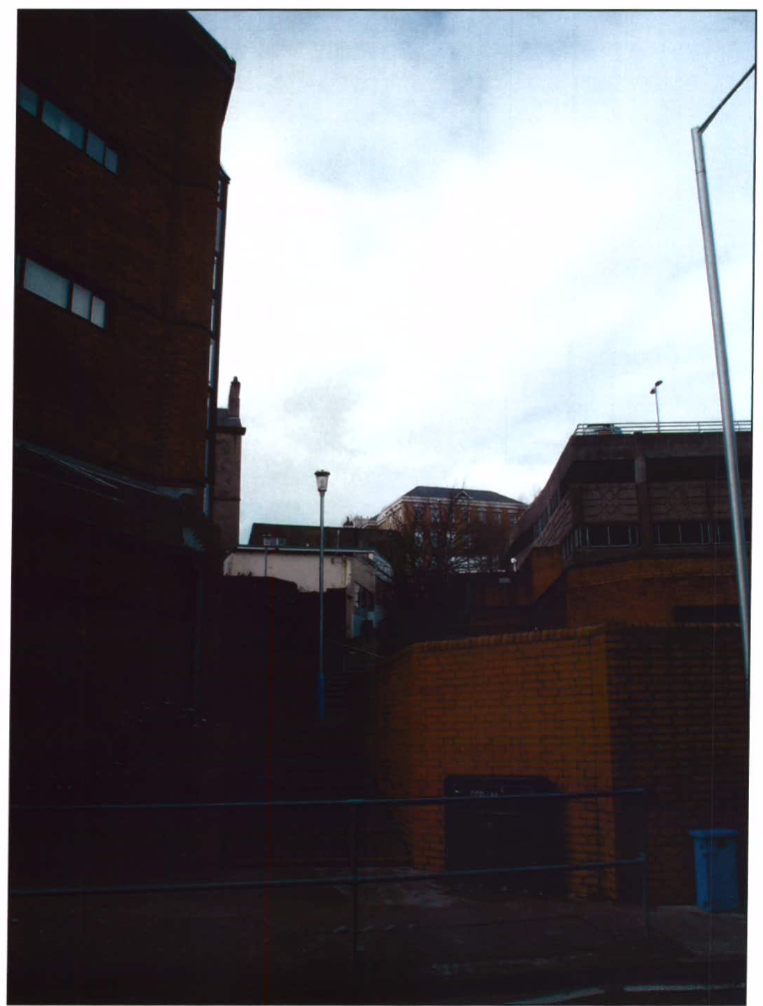 A photograph showing the existing urban surroundings of the site, featuring brick buildings, a retaining wall, and a street lamp.