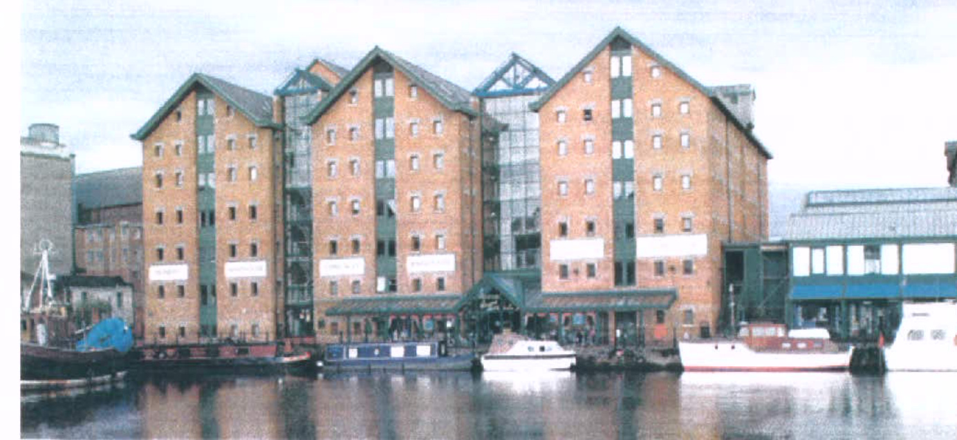 A photograph showing a large, multi-story brick building complex situated along a waterfront with boats moored in the foreground.