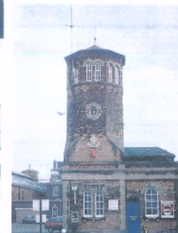 A grainy photograph showing a historic stone tower structure with arched windows and a crest, attached to a lower building.